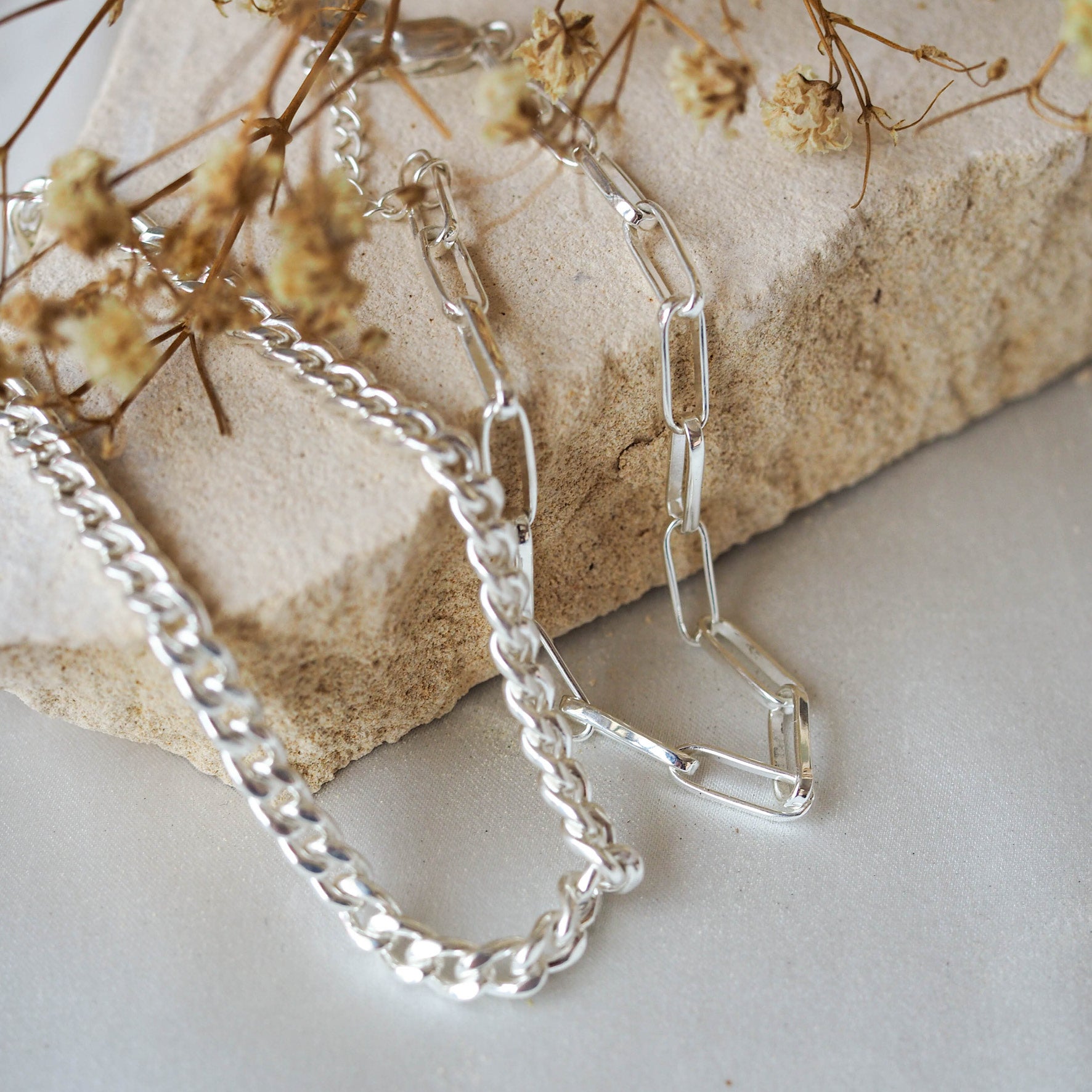 Two silver chain bracelets with different link styles displayed on a textured beige stone with dried flowers in the corner