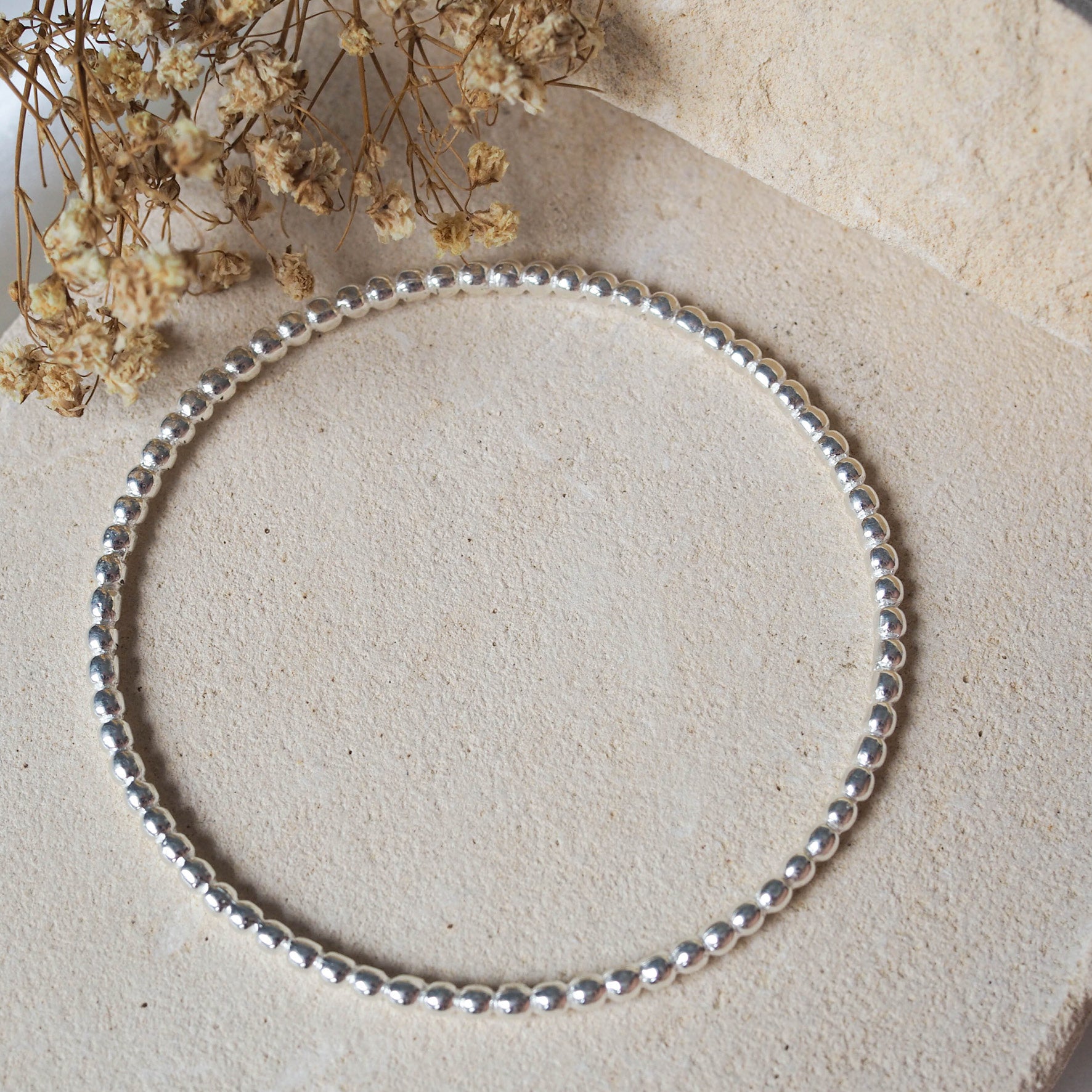 A silver beaded bracelet laid flat on a textured beige stone with dried flowers arranged nearby.