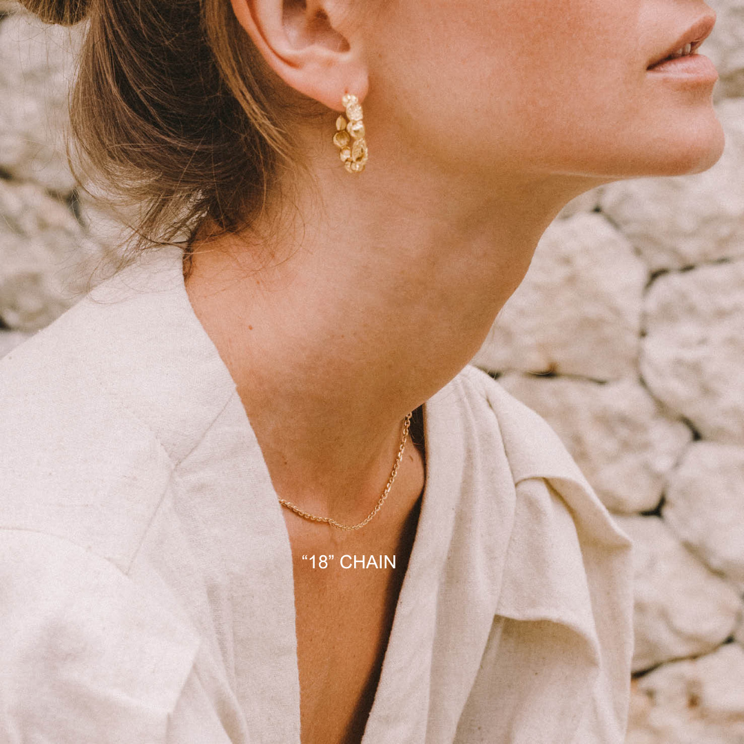 Close-up of a person wearing gold earrings and a necklace with a stone wall background