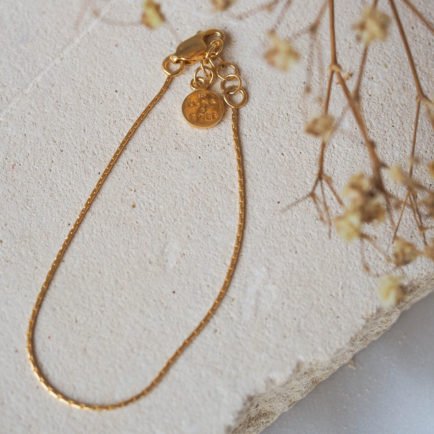 A delicate gold chain bracelet, displayed on beige stone with dried flowers.