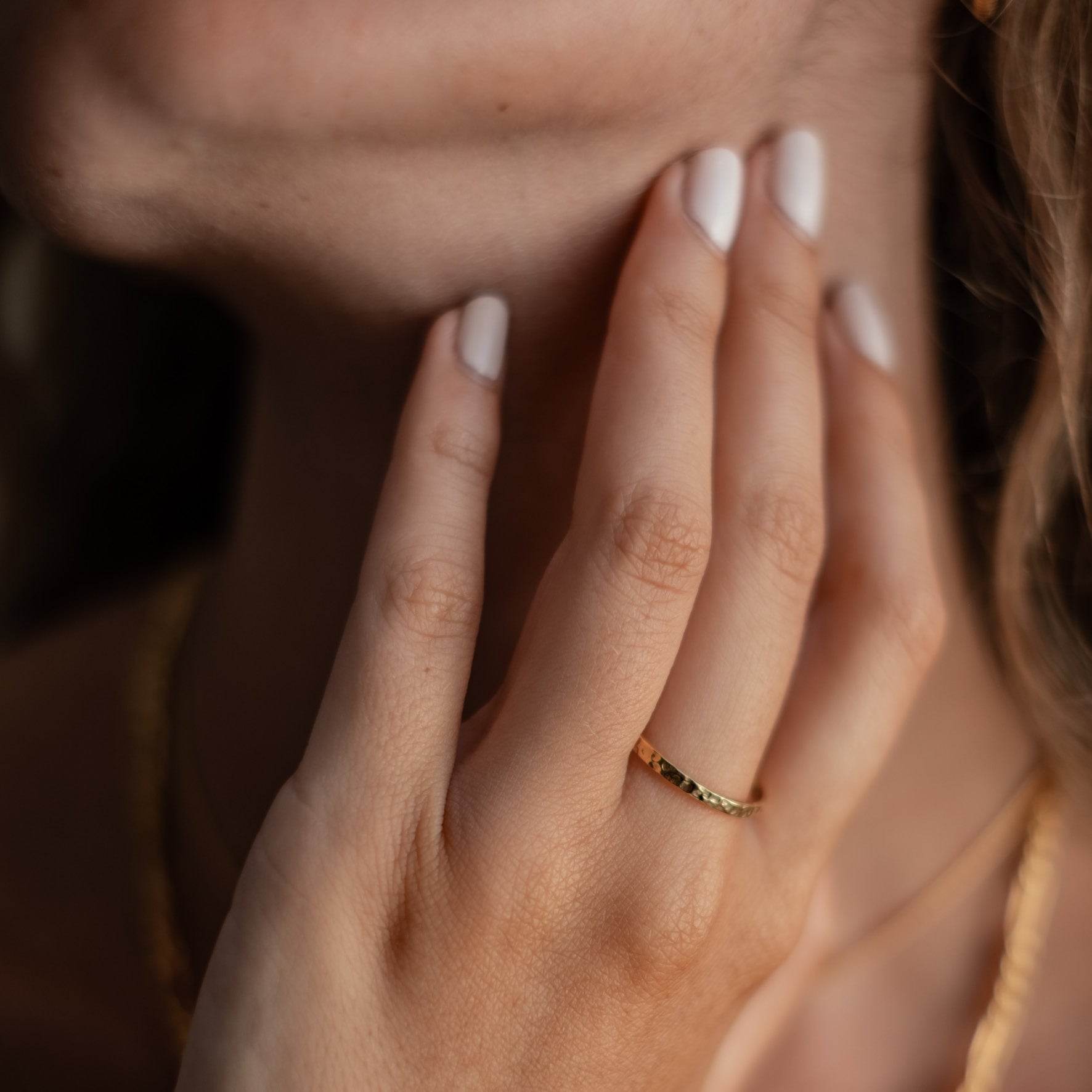 A close-up of a woman's hand with white nail polish touching her neck, wearing a thin gold ring.
