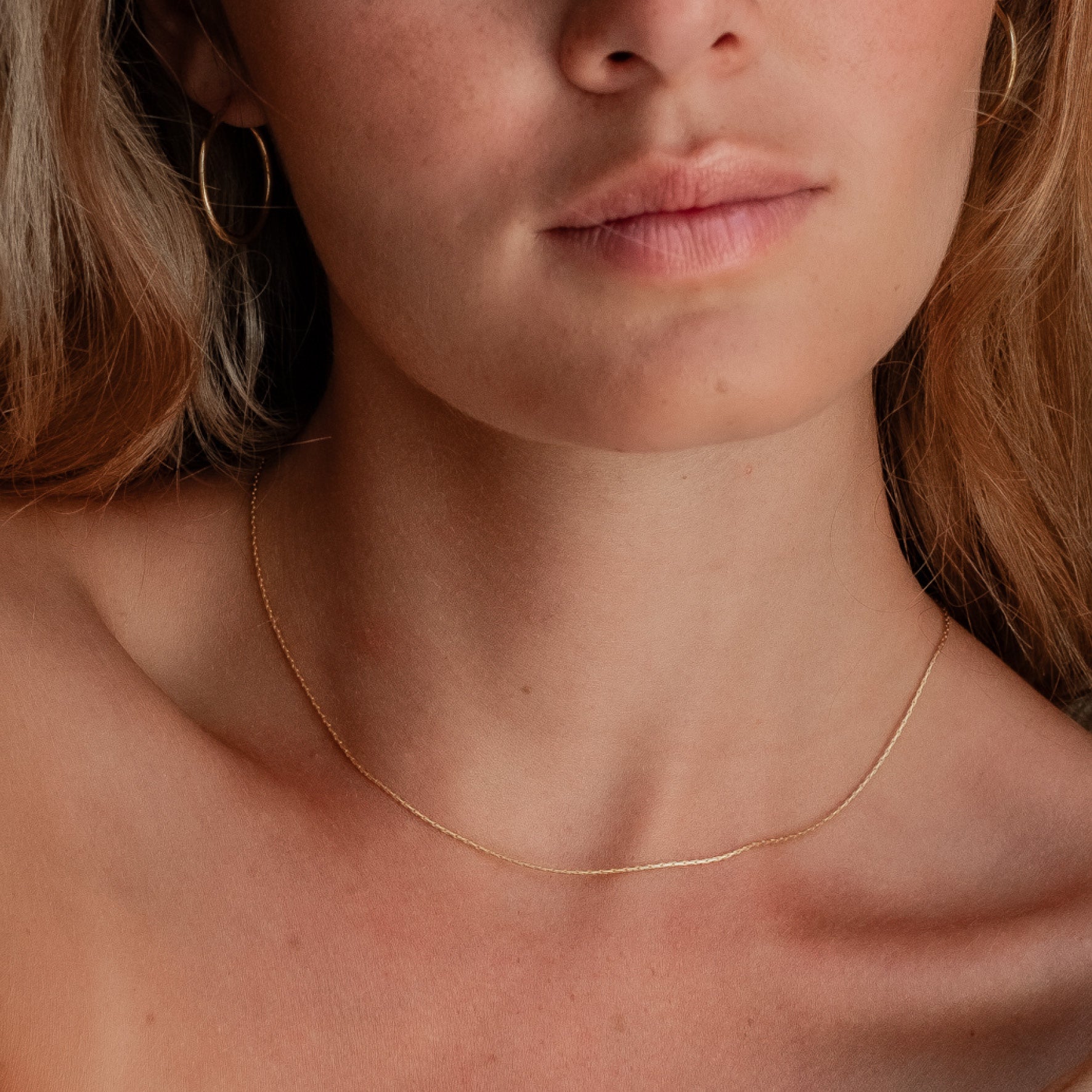 Close-up of a woman's neck and shoulders wearing a thin gold chain necklace and gold hoop earring.