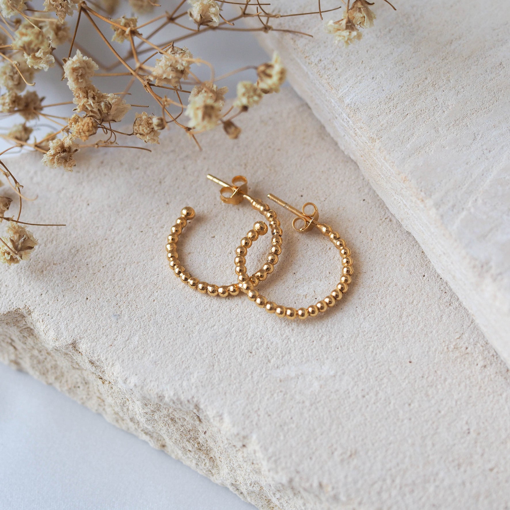 Gold beaded earrings displayed on a textured beige stone with dried flowers.