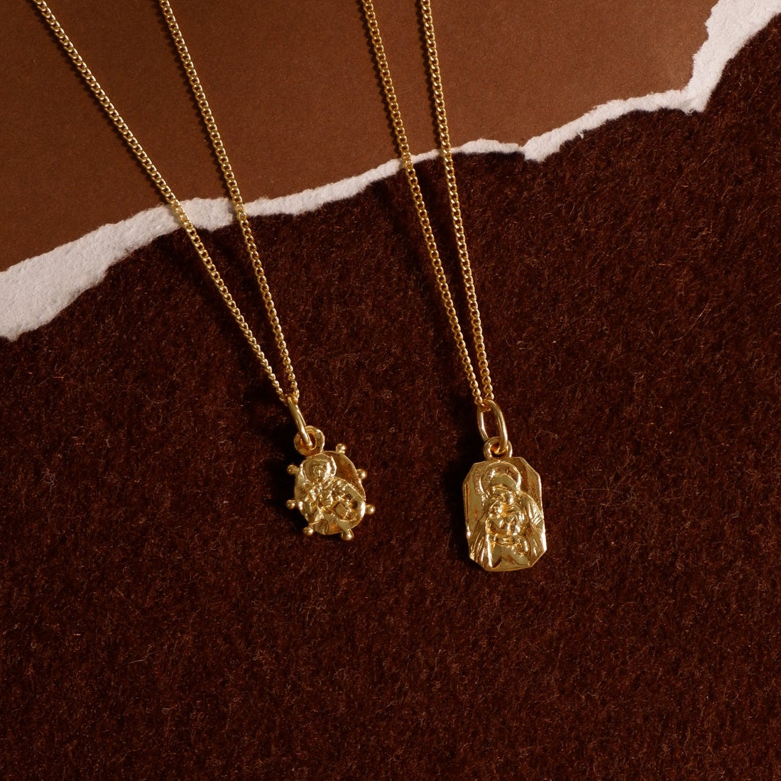 Two gold Saint necklaces with unique pendants on a brown & red textured background