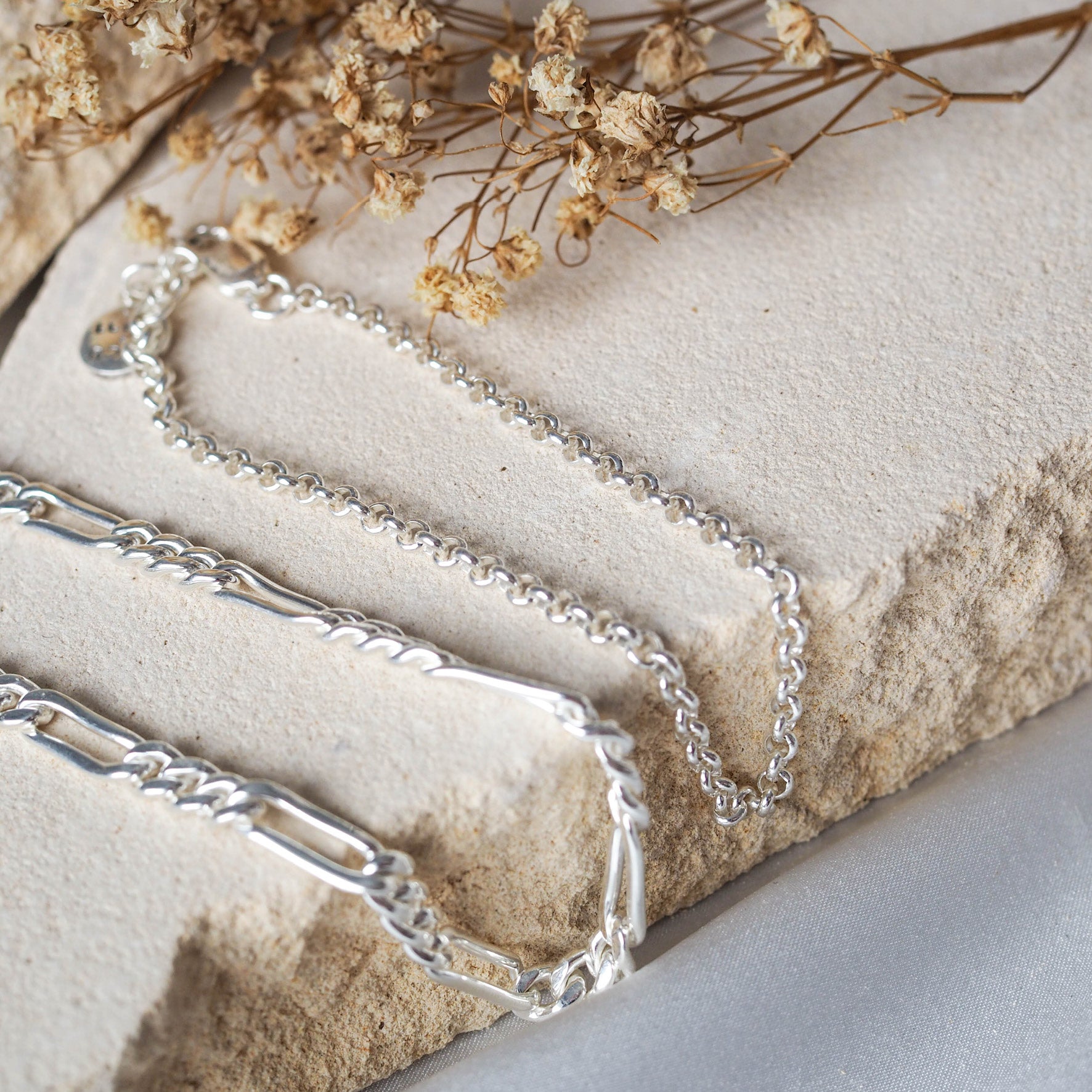 A silver chain bracelet with an adjustable clasp and circular charm, displayed on beige stone with dried flowers.