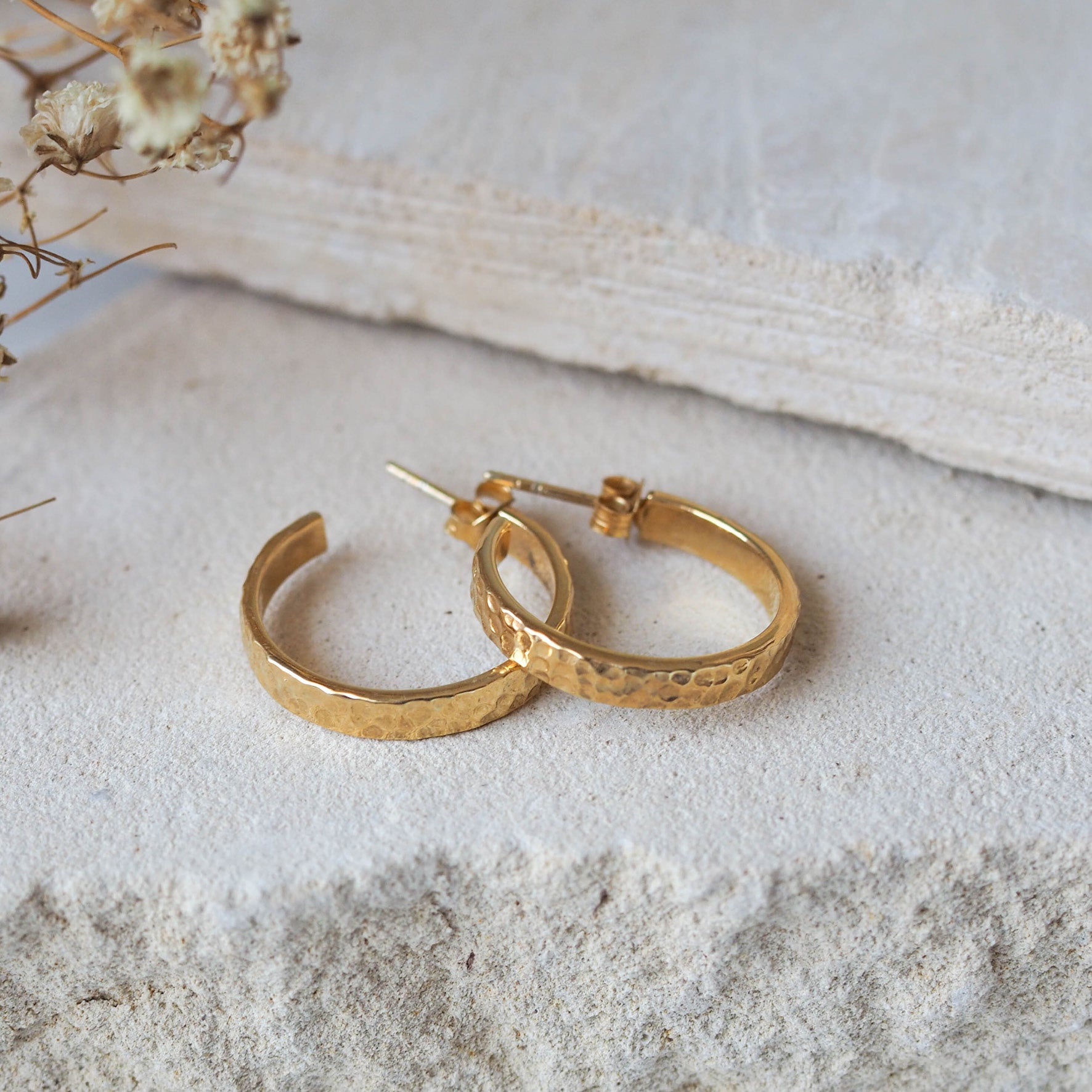A pair of hammered gold hoop earrings displayed on textured stone beside dried flowers.