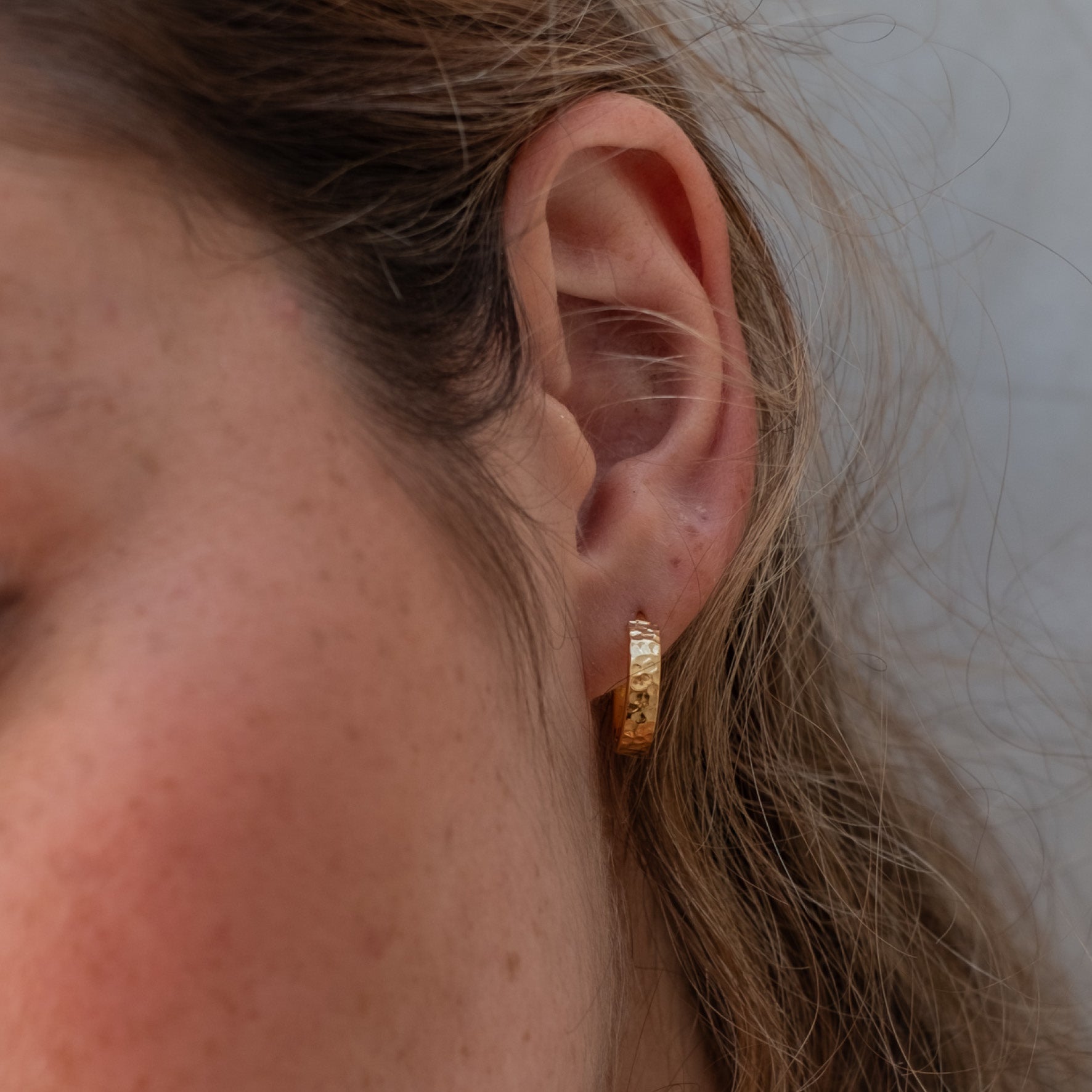 Close-up of a single hammered gold hoop earring worn on an ear with light brown hair.