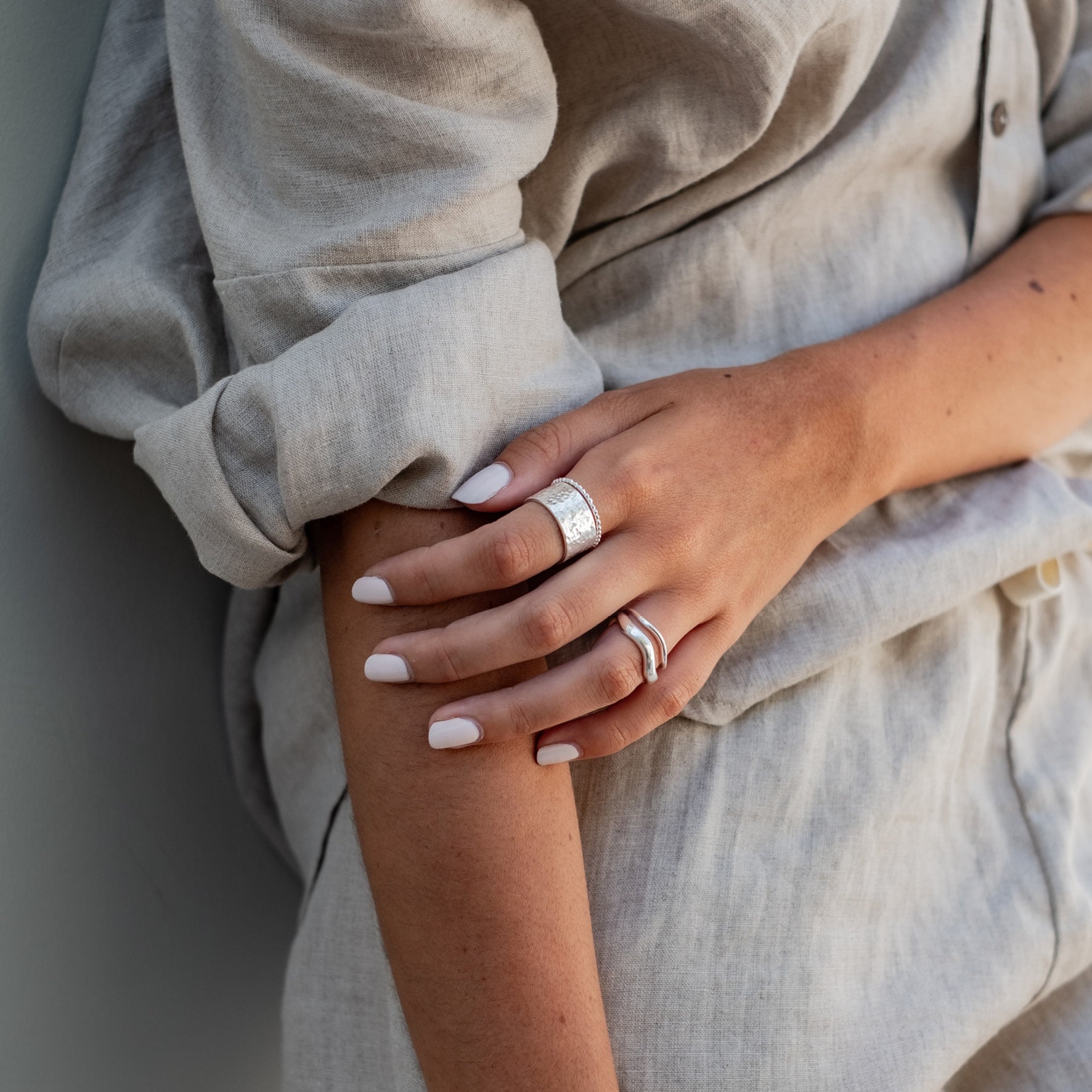 A close-up of a woman wearing two silver rings on their fingers, with their hand resting on their arm, dressed in a light beige outfit.