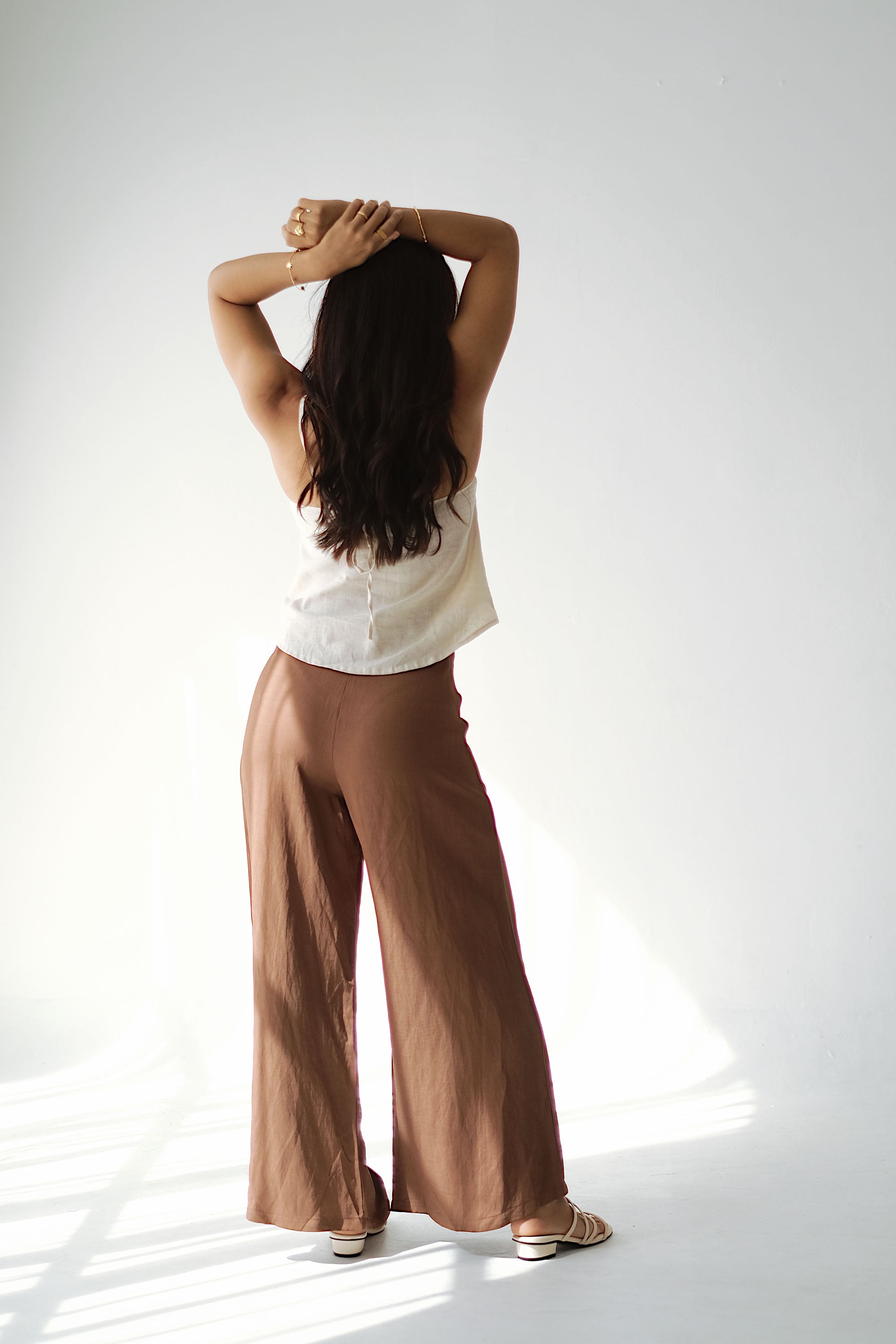 Person wearing a white top and brown pants standing with hands behind head on a white background