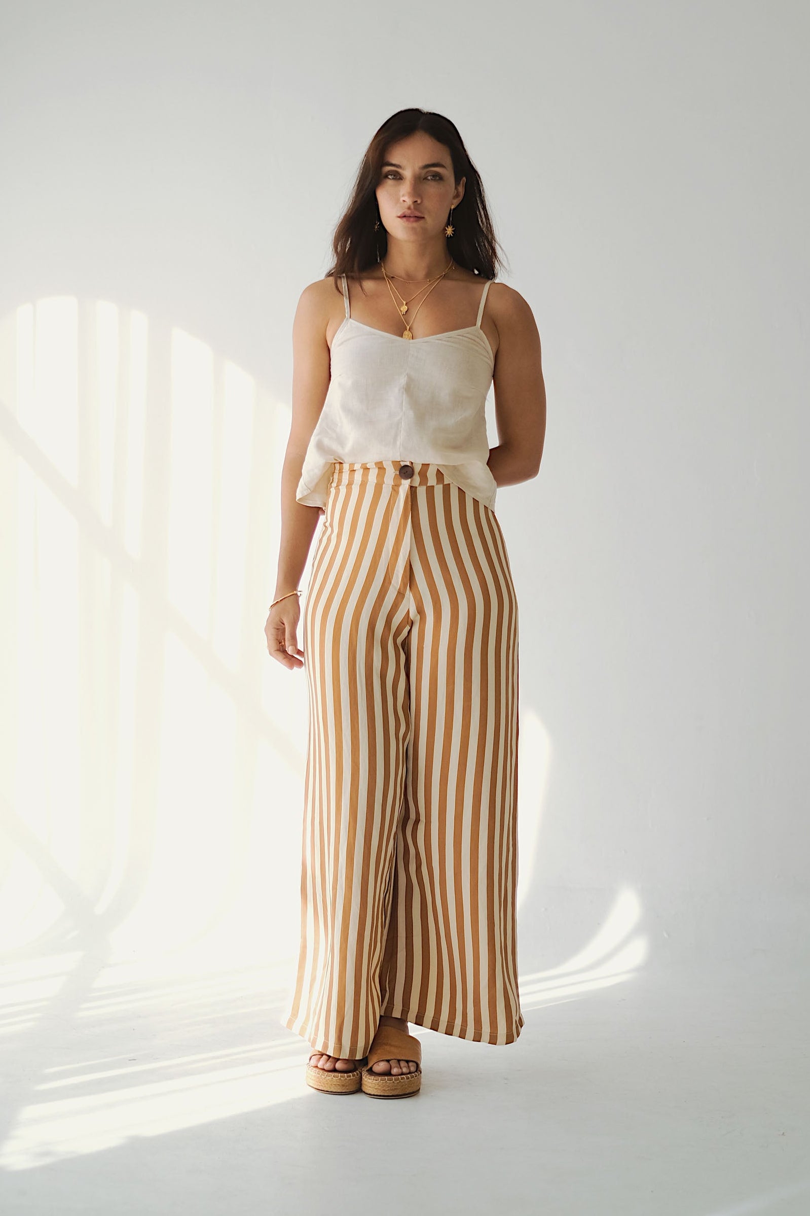 Woman wearing a white top and striped pants standing in a well-lit room with sunlight filtering through curtains.
