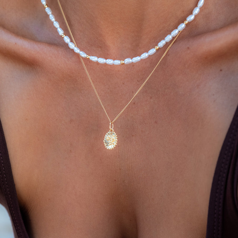 A close-up of a model's chest wearing two layered necklaces: a short white pearl strand and a gold chain with a textured oval pendant.