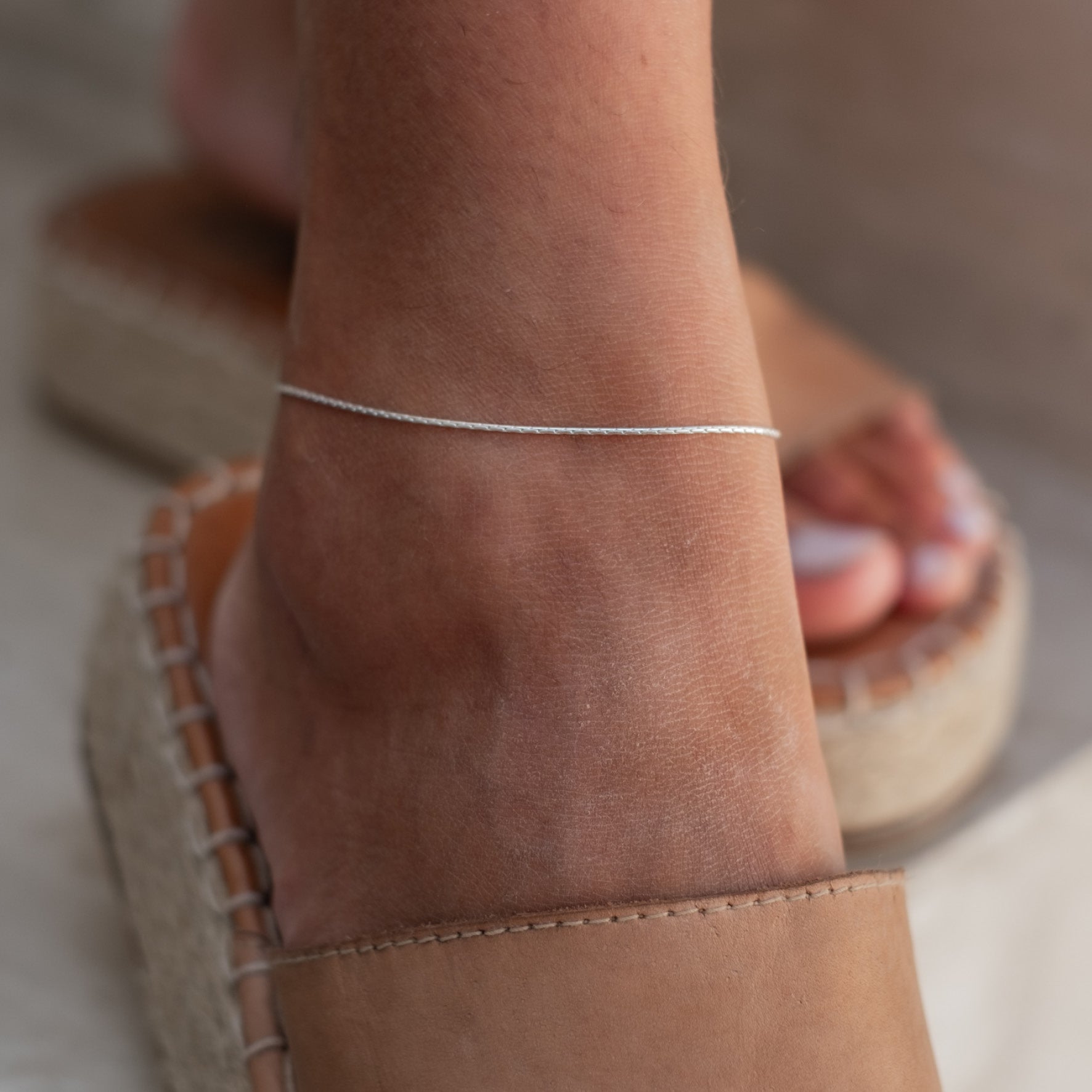 Close-up of a woman's ankle wearing a delicate silver anklet, paired with beige espadrilles.