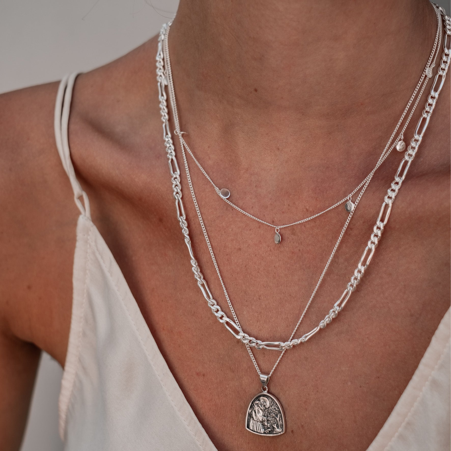 A close-up of a person wearing layered silver necklaces, including a medallion pendant and delicate chains, shown from the shoulders up.