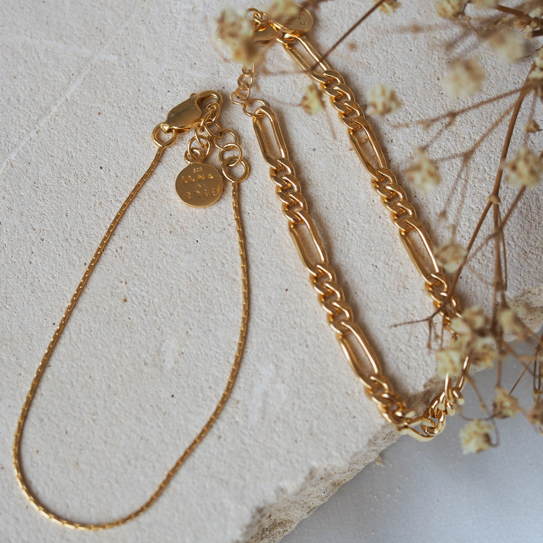 Two gold chain bracelets, one with elongated links and the other a delicate chain, displayed on beige stone with dried flowers.