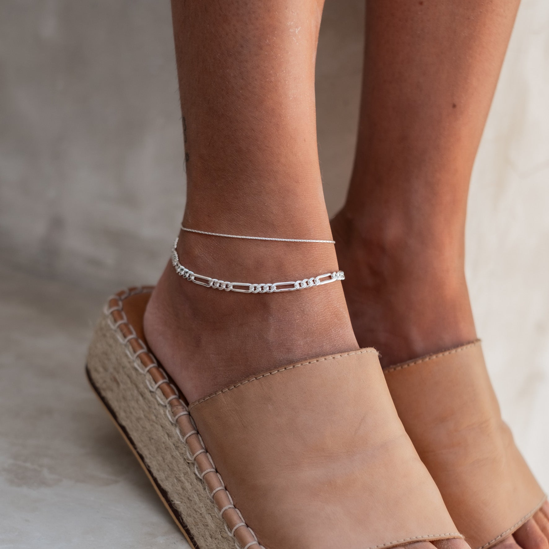 Close-up of a woman's ankle wearing a delicate silver anklet and one with links, paired with beige espadrilles.