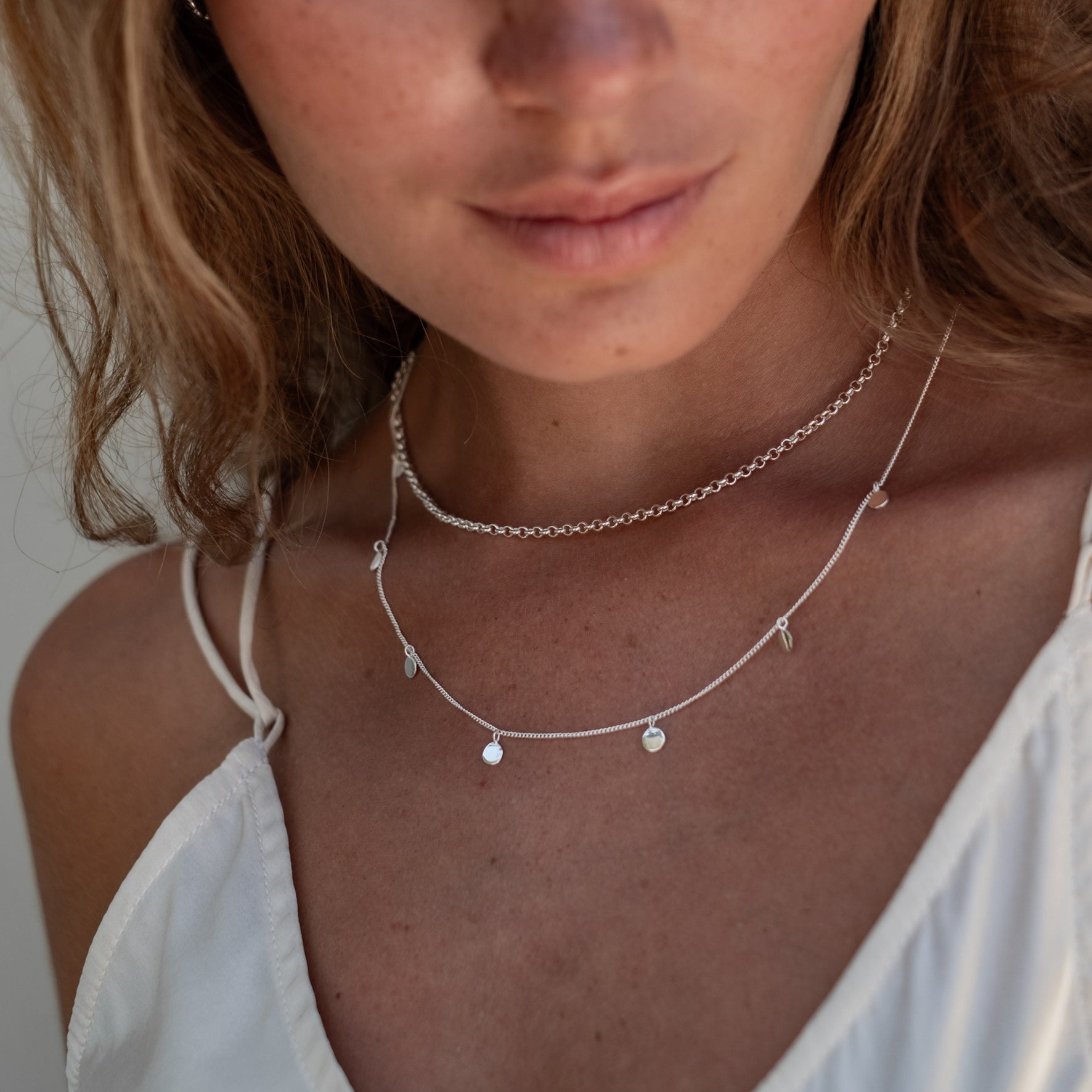 Close-up some silver necklaces on a woman's neck, who is wearing a white strappy top.