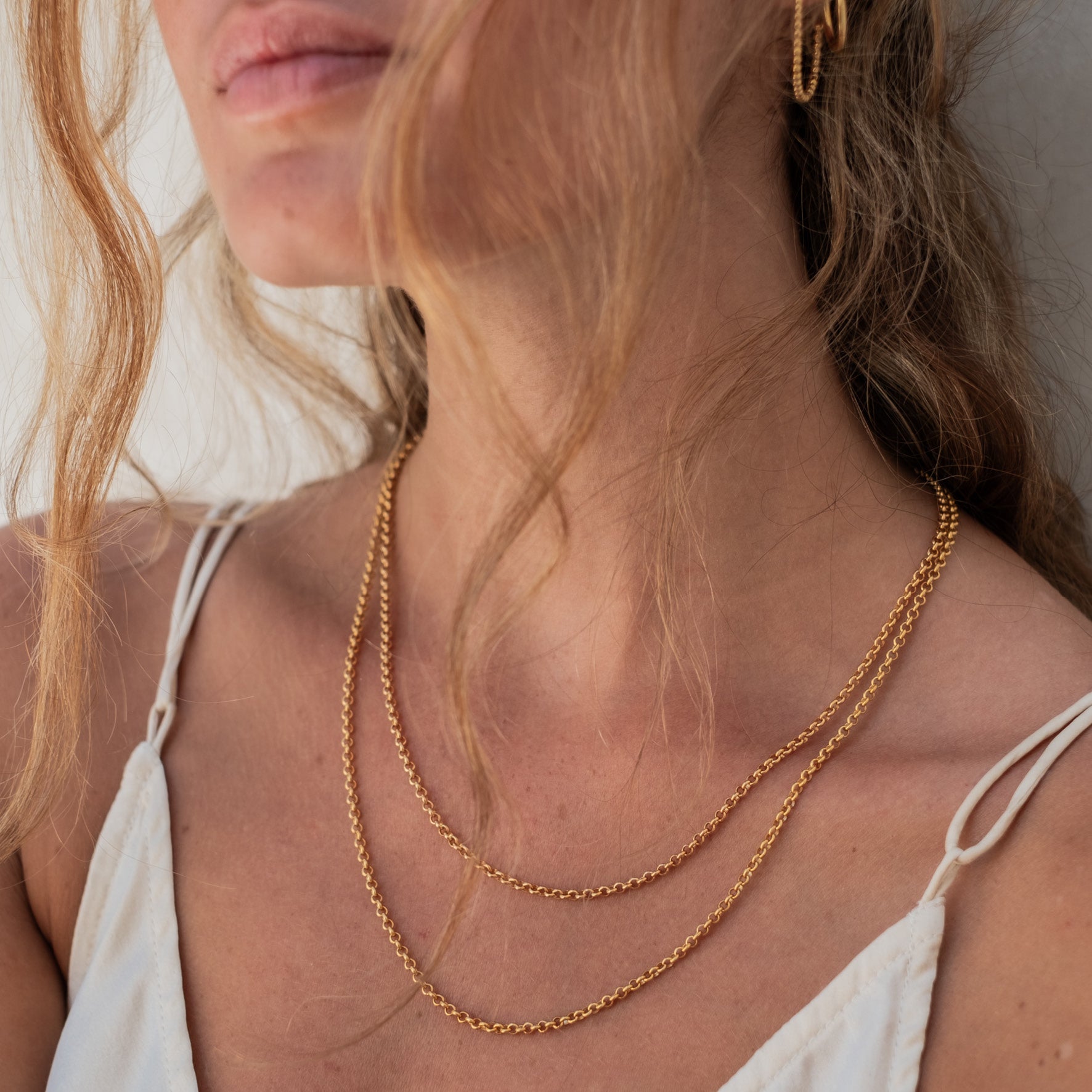 Close-up of a woman's neck and shoulders wearing a thin golden chain necklaces and golden hoop earring.