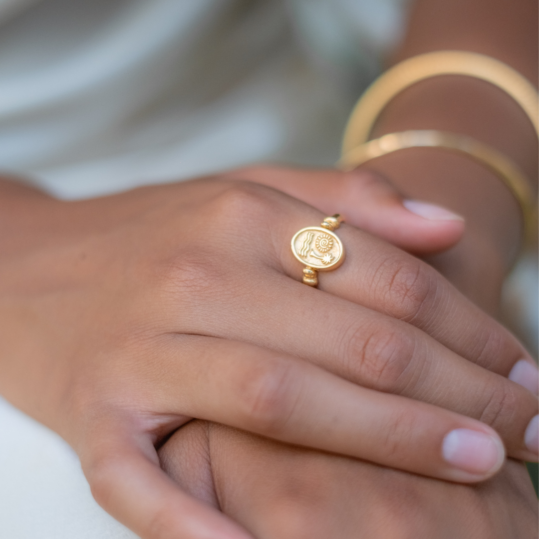A close-up of a gold ring with a sun and waves design on a woman's finger, alongside a gold bracelet.