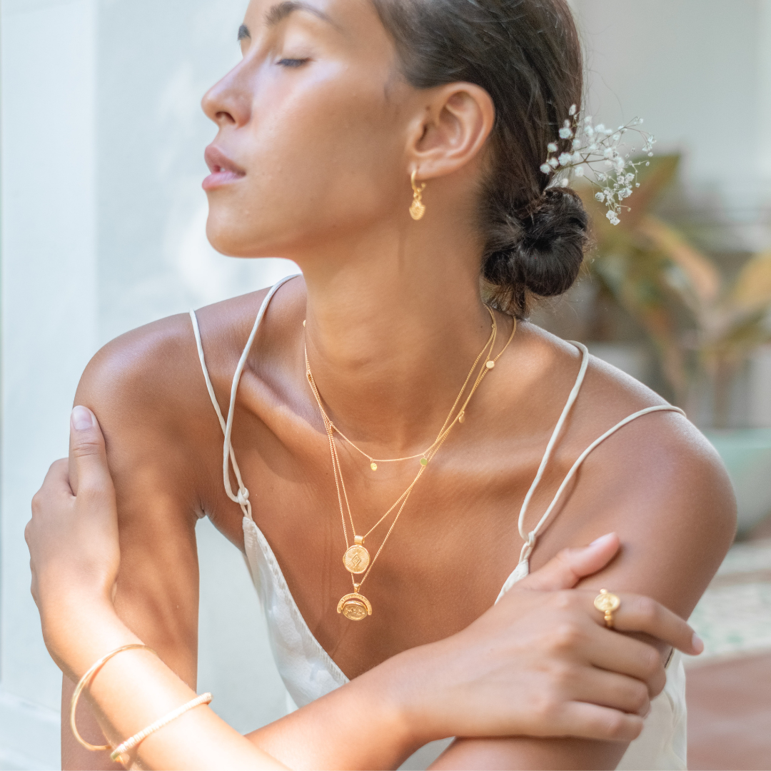 A woman wearing layered gold necklaces with matching earrings, rings, and a bracelet, dressed in a white top and seated in an elegant setting.