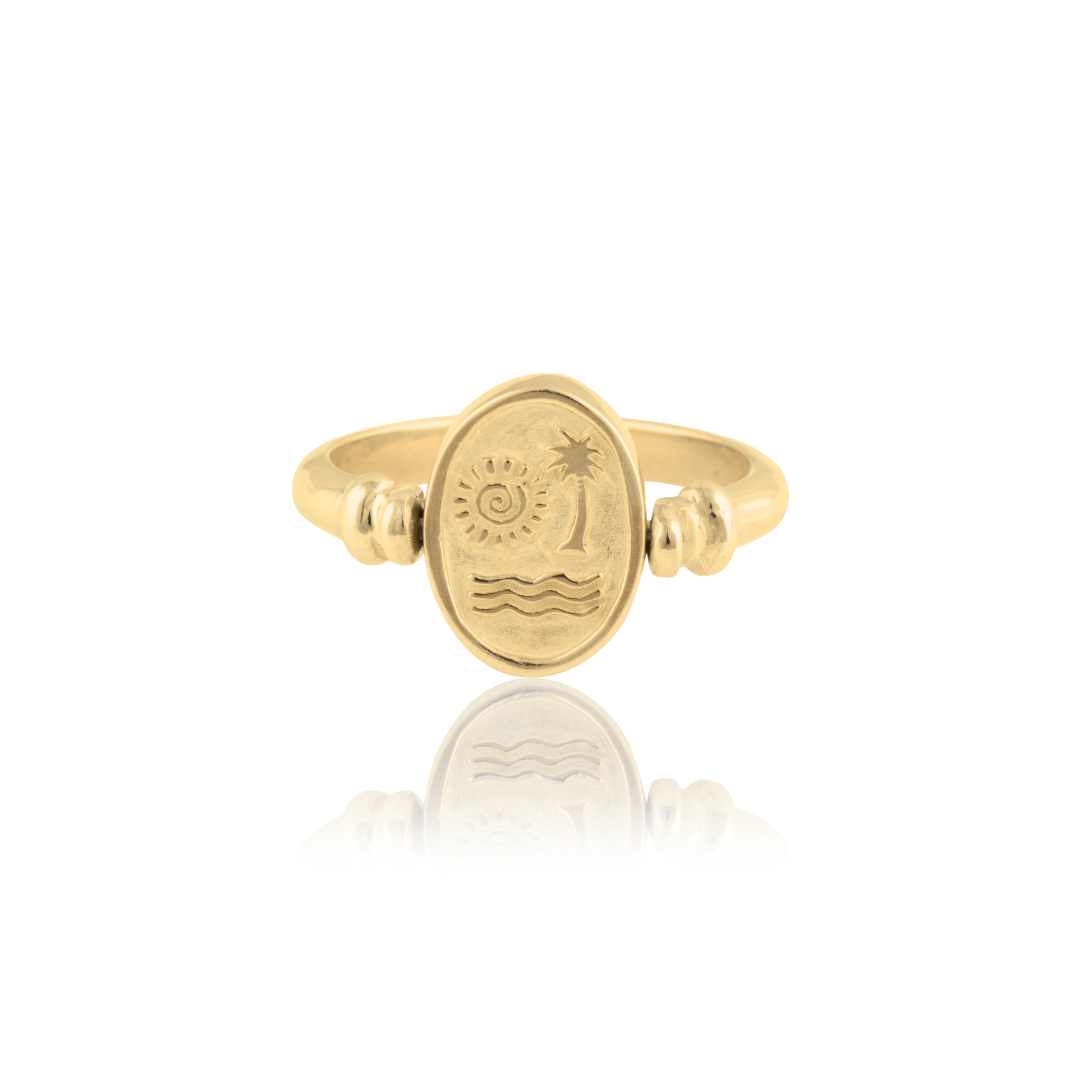 A gold ring with an oval face depicting a sun, palm tree, and waves, reflected on a glossy surface.