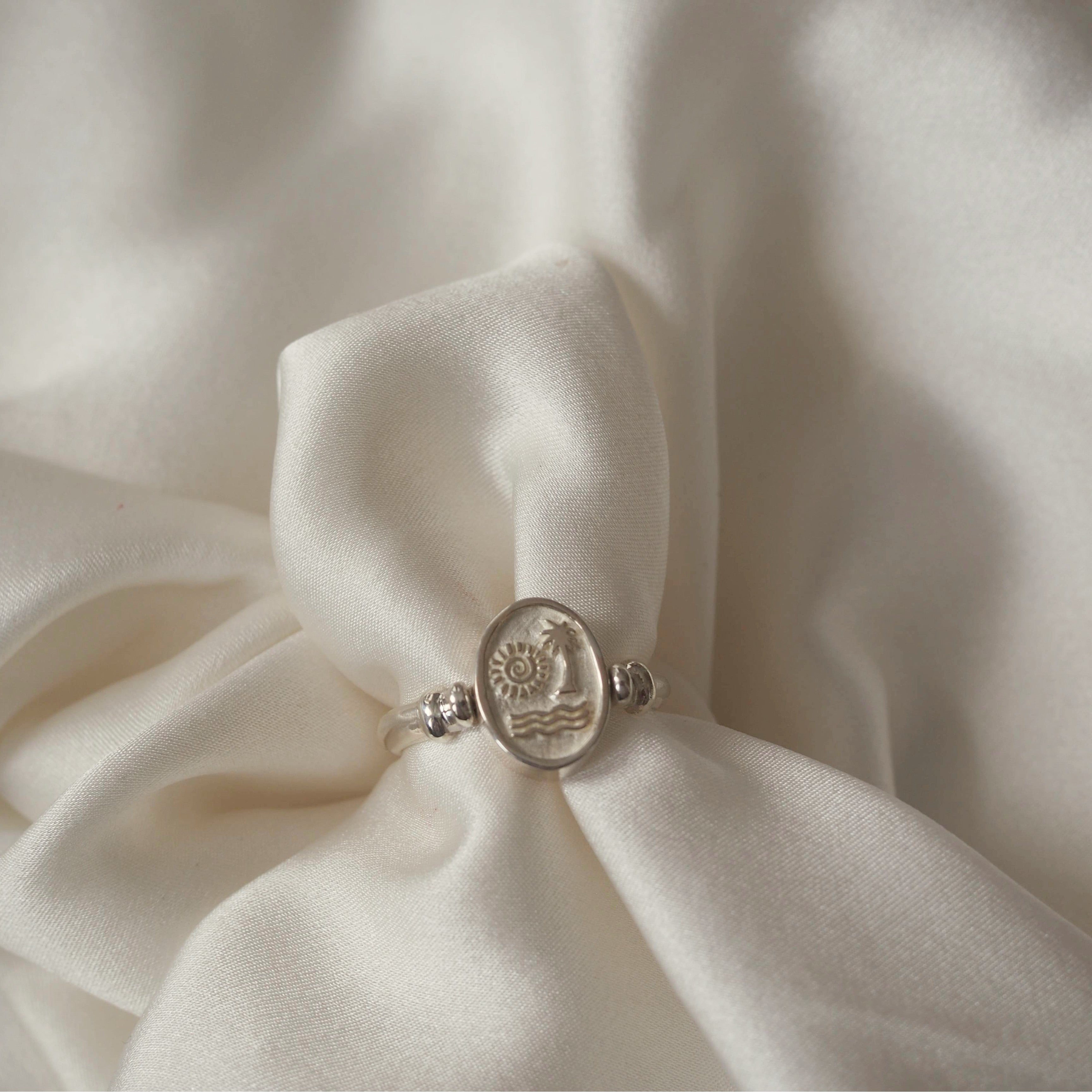 A silver ring with an oval face depicting a sun, palm tree, and waves design, elegantly displayed on a gathered white satin fabric.