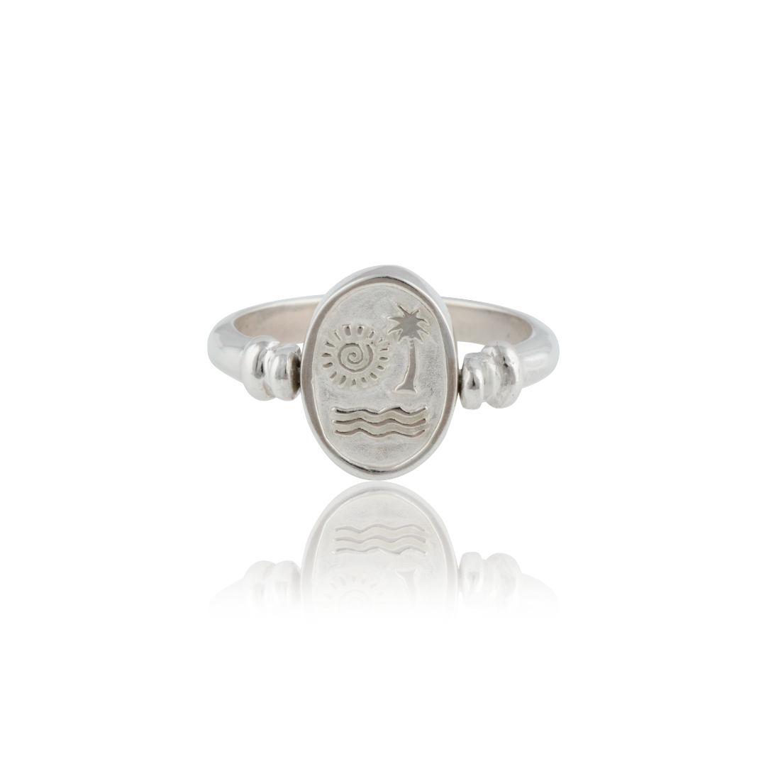 A silver ring with an oval face depicting a sun, palm tree, and waves, reflected on a glossy surface.