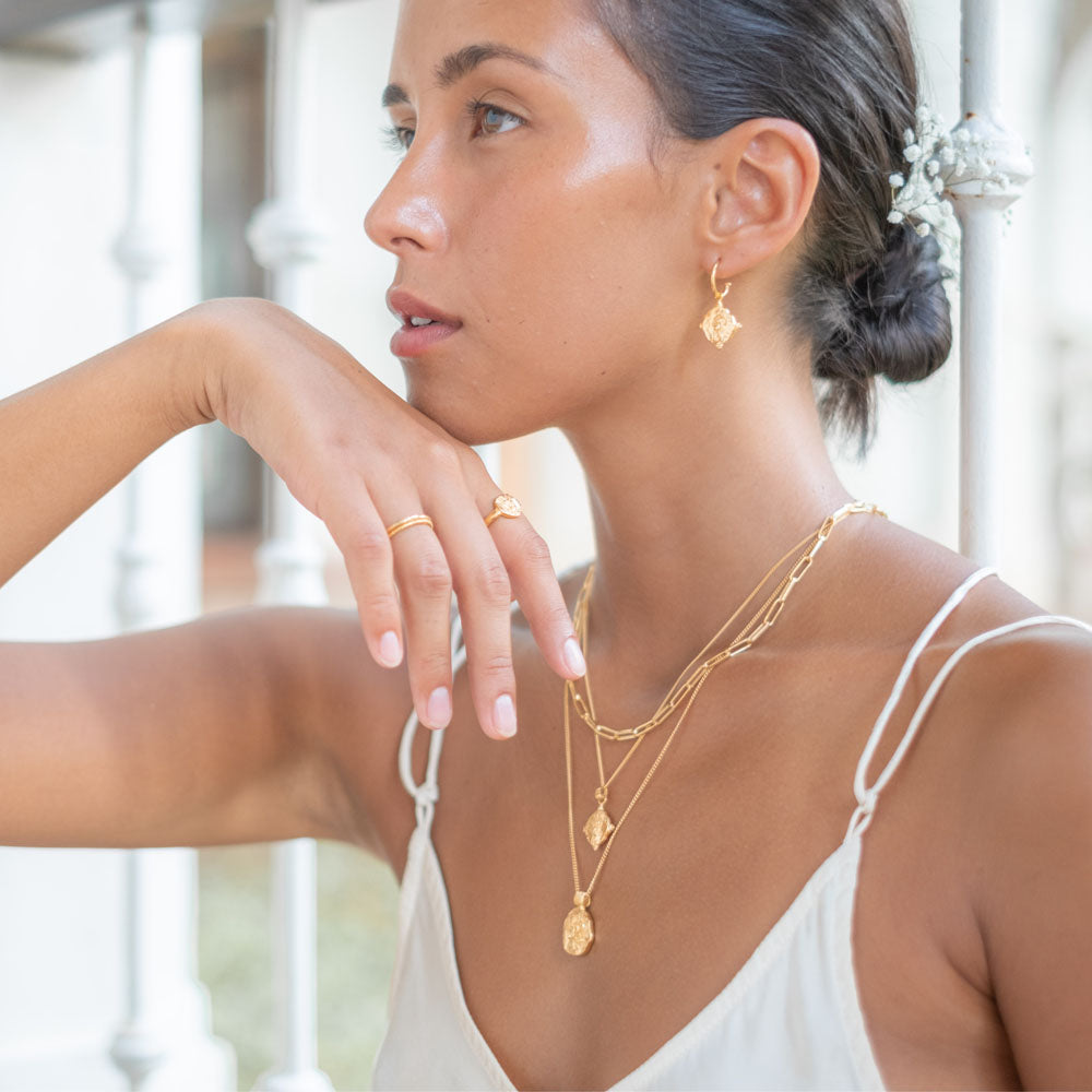 A woman wears a gold hoop earring and a multiple gold necklaces, with her hair styled in a low bun adorned with small white flowers."
