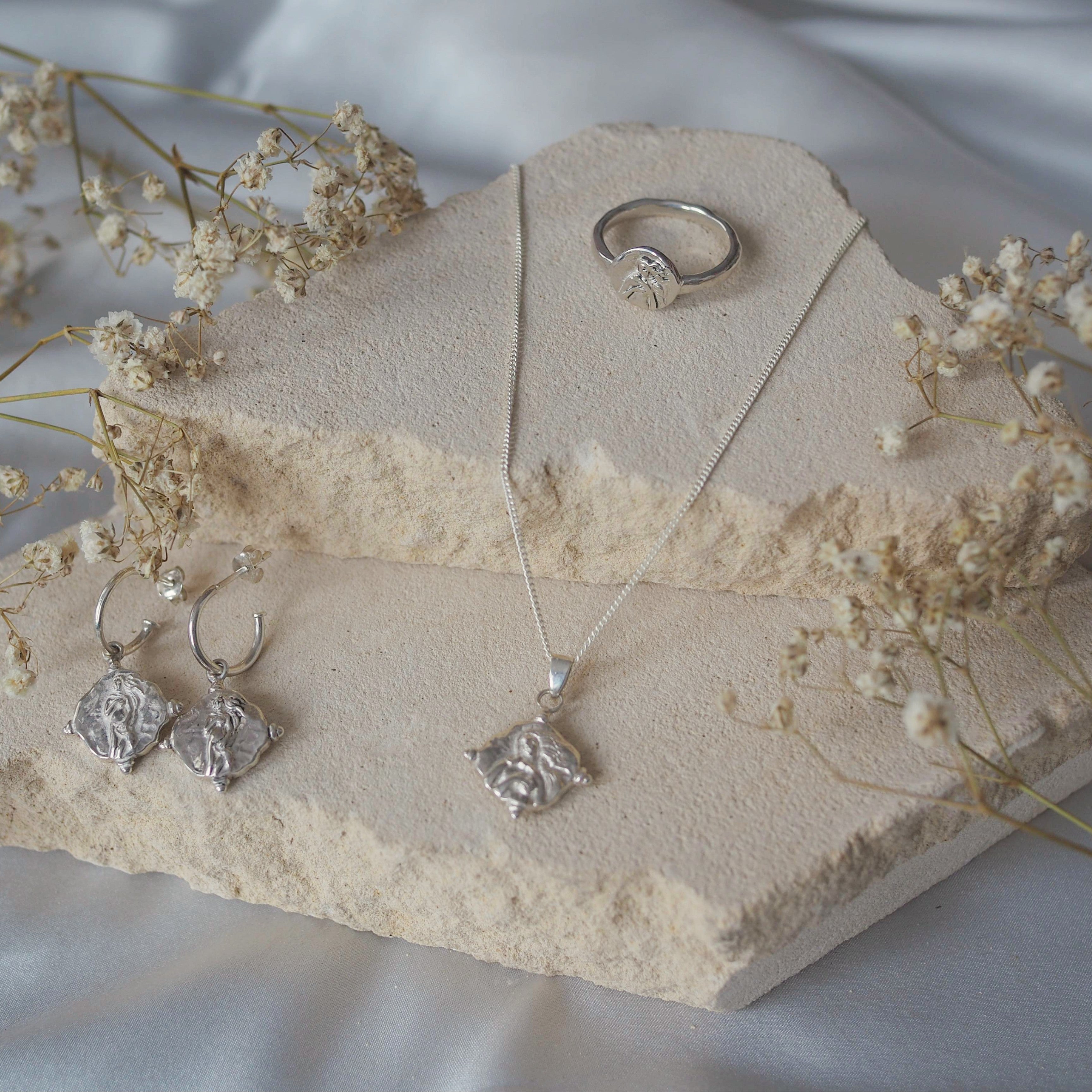 A silver jewelry set, including a pair of hoop earrings with religious figure design charms, a necklace and a ring with a similar design, is displayed on a beige stone with delicate white flowers surrounding it.