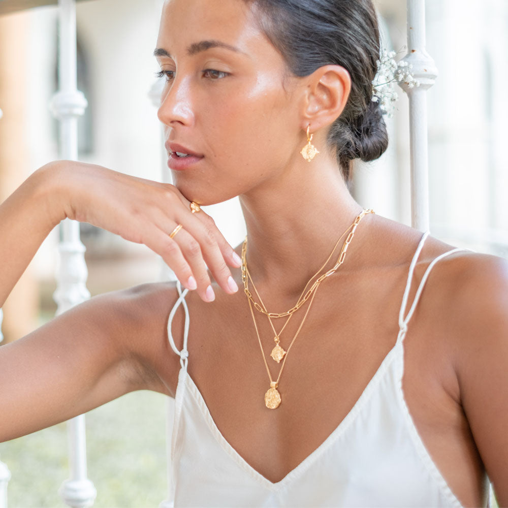 A woman wears a gold hoop earring and a multiple gold necklaces, with her hair styled in a low bun adorned with small white flowers.
