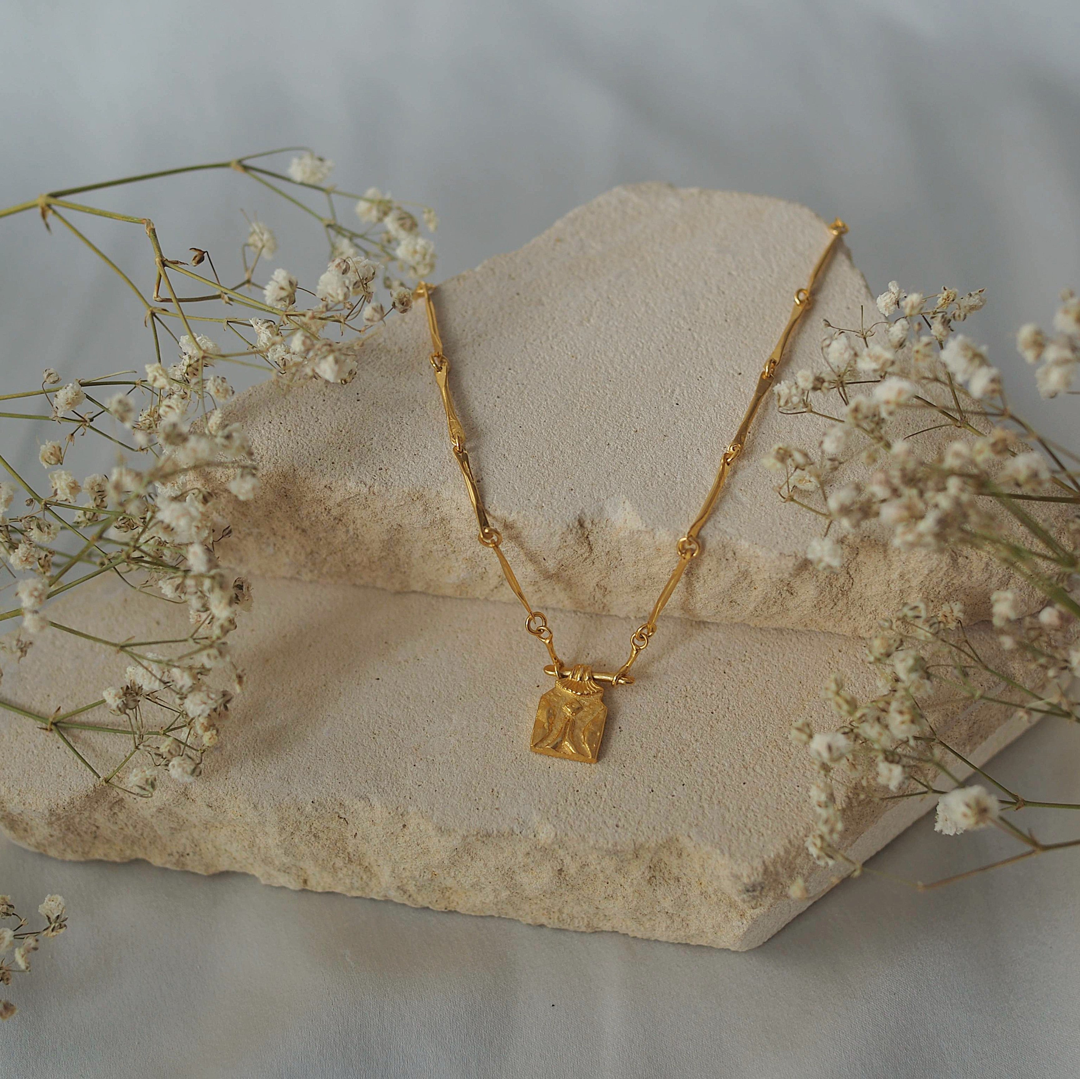 A gold necklace, displayed on a light stone surface with some dried plants in the background.