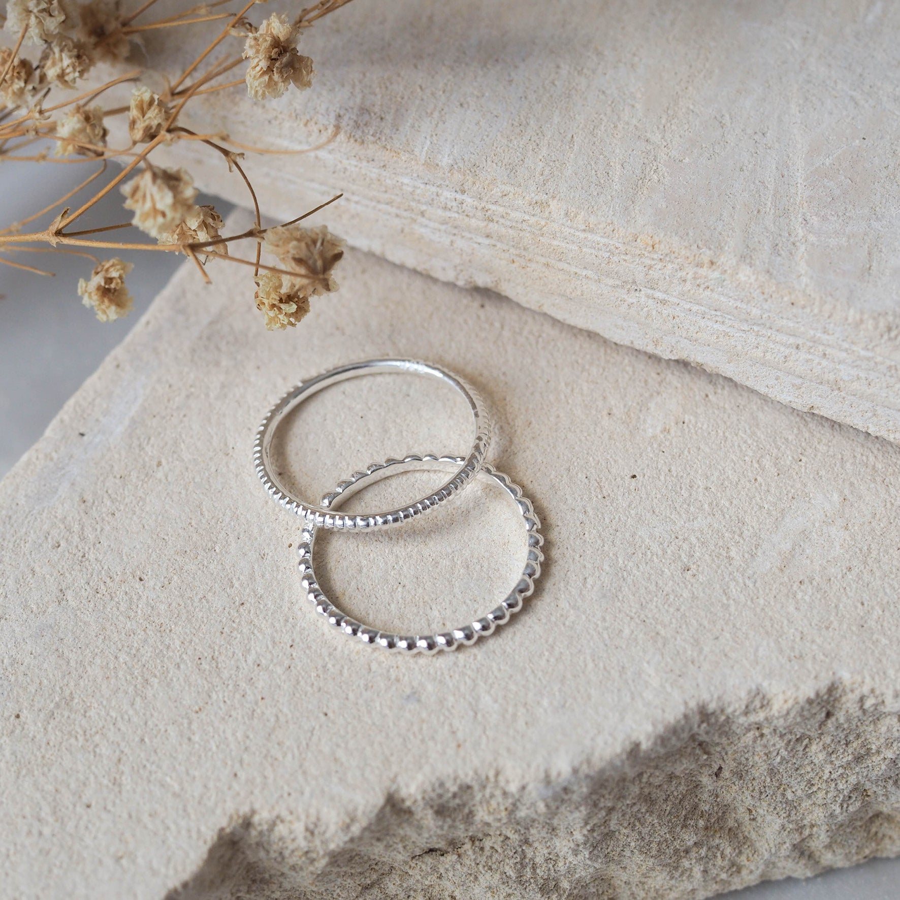 Silver beaded rings displayed on a textured beige stone with dried flowers.