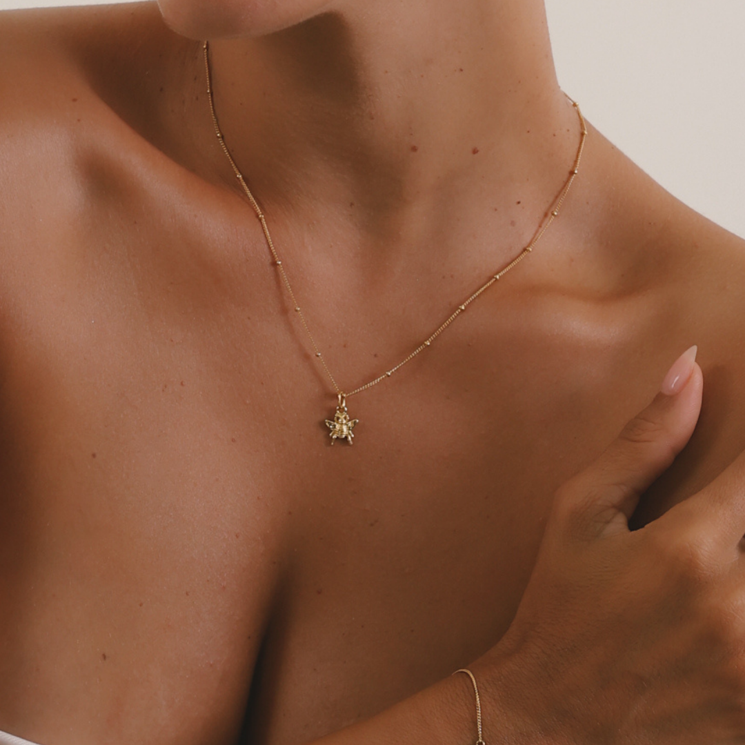 A delicate gold chain necklace with a small flower-like pendant, worn on a model's neck. The shot is a close-up lifestyle crop focusing on the jewelry against the model's skin.