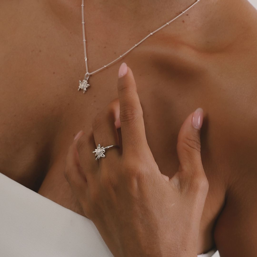 Close-up of a necklace and ring with a hand touching the chest, on a neutral background.