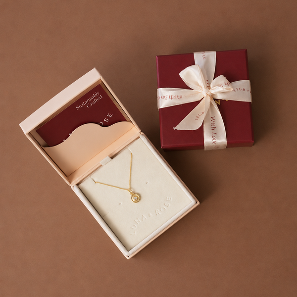 A gold necklace with a small circular pendant displayed inside an open cream jewelry box next to a dark red gift box tied with a cream ribbon, on a solid brown background.