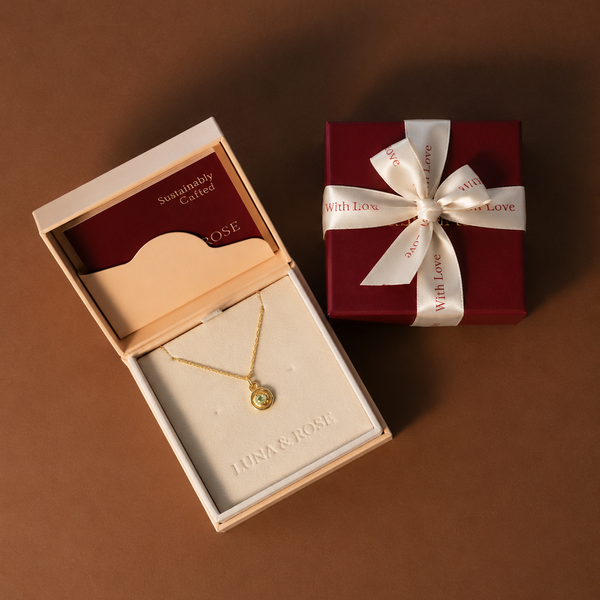 A gold necklace with a small round pendant displayed in an open cream jewelry box, alongside a dark red gift box tied with a creme ribbon