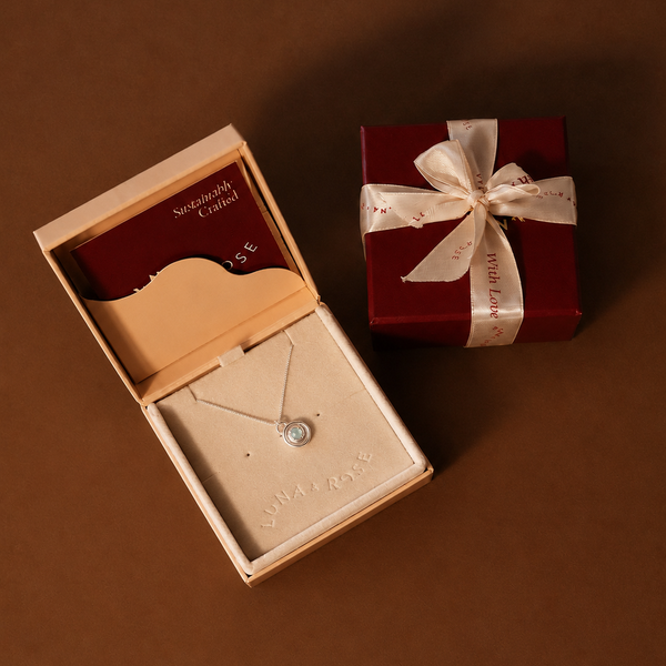 A silver necklace with a round clear stone displayed in an open beige jewelry box, alongside a dark red gift box tied with a cream ribbon on a solid brown background.