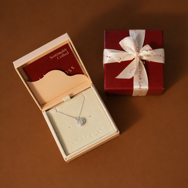 A silver necklace with a round clear stone displayed in an open cream-colored jewelry box, alongside a red gift box tied with a white ribbon on a brown background.
