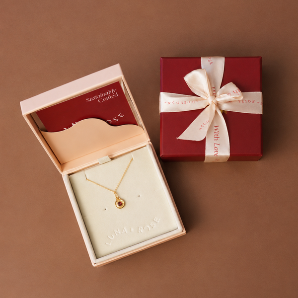 A gold necklace with a small round pendant displayed in an open cream jewelry box, alongside a closed dark red gift box tied with a cream ribbon on a brown background.