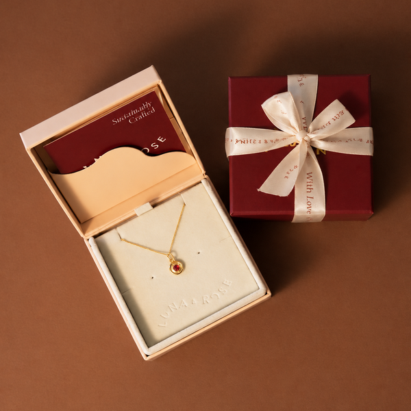 A gold necklace with a small red oval gemstone displayed in an open cream jewelry box, alongside a dark red gift box tied with a cream ribbon and bow.