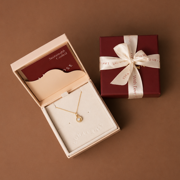 A gold necklace with a small round pendant displayed in an open cream jewelry box, alongside a dark red gift box tied with a cream ribbon