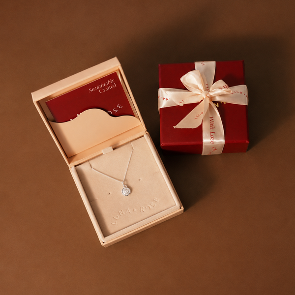 A silver necklace with a small round stone displayed in an open cream jewelry box, alongside a red gift box tied with a white ribbon on a brown background.