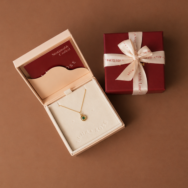 A gold necklace with a small green oval stone pendant is displayed inside an open cream-colored jewelry box, next to a closed dark red gift box tied with a cream ribbon
