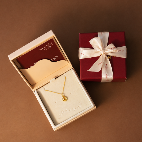 A gold necklace with a small round pendant displayed in an open cream jewelry box, alongside a wrapped red gift box tied with a white ribbon on a brown background.