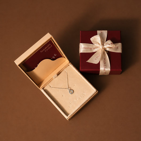 A silver pendant necklace displayed in an open beige jewelry box next to a dark red gift box tied with a cream ribbon, on a solid brown background.