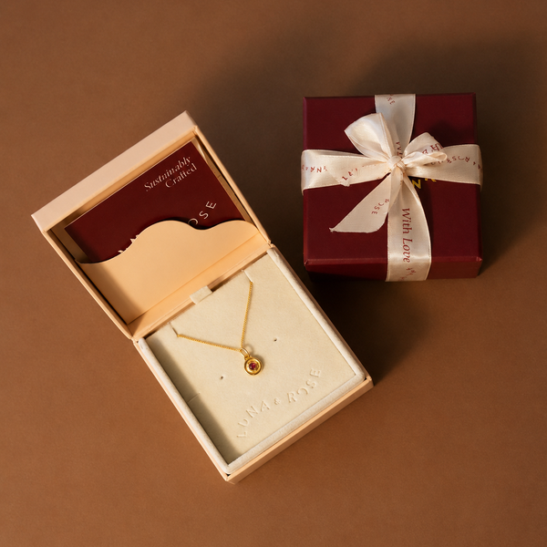 A gold necklace with a small round pendant displayed in an open cream jewelry box, alongside a dark red gift box tied with a white ribbon on a solid brown background.