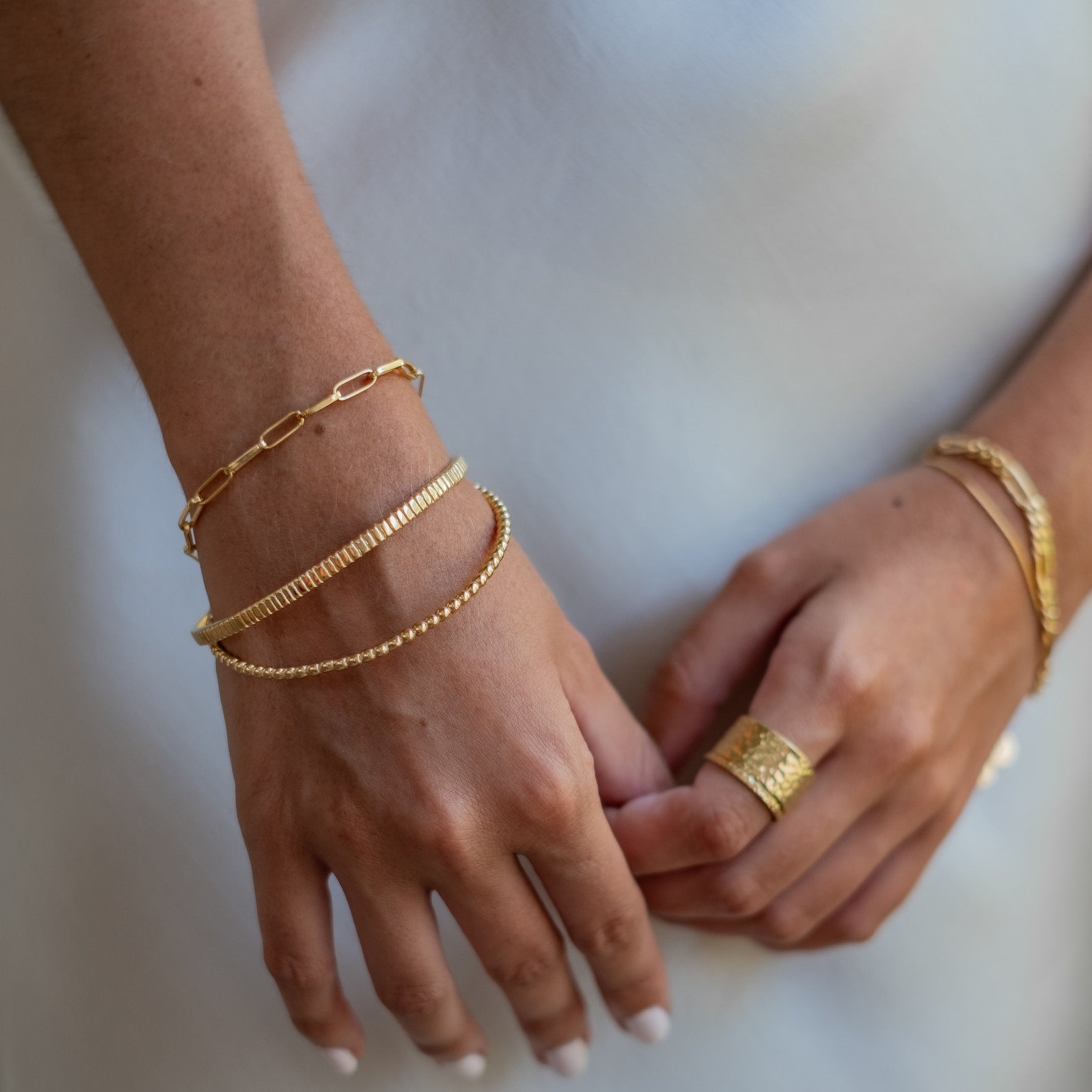 Multiple gold bracelets, including a beaded one, worn on both wrists along with a gold ring on a finger, against a light background.