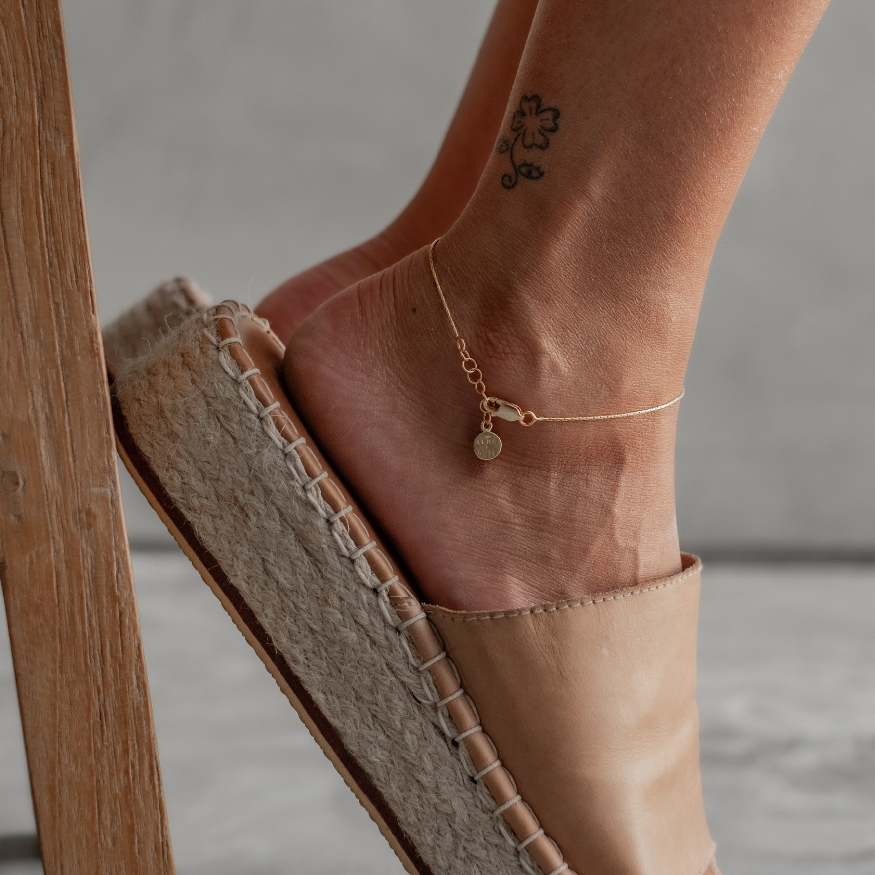 Close-up of a woman's ankle wearing a delicate gold anklet, paired with beige espadrilles.