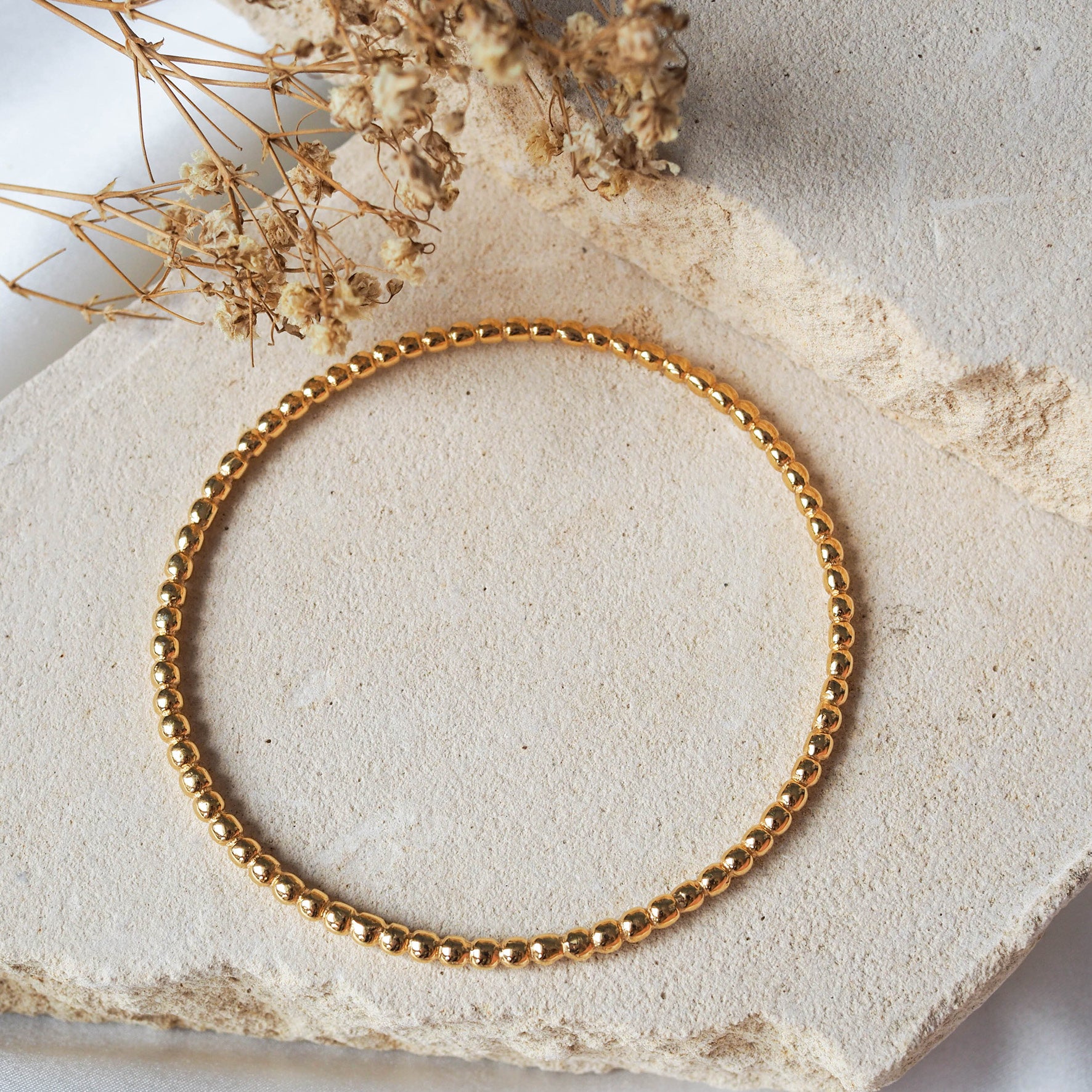 A gold beaded bracelet laid flat on a textured beige stone with dried flowers arranged nearby.
