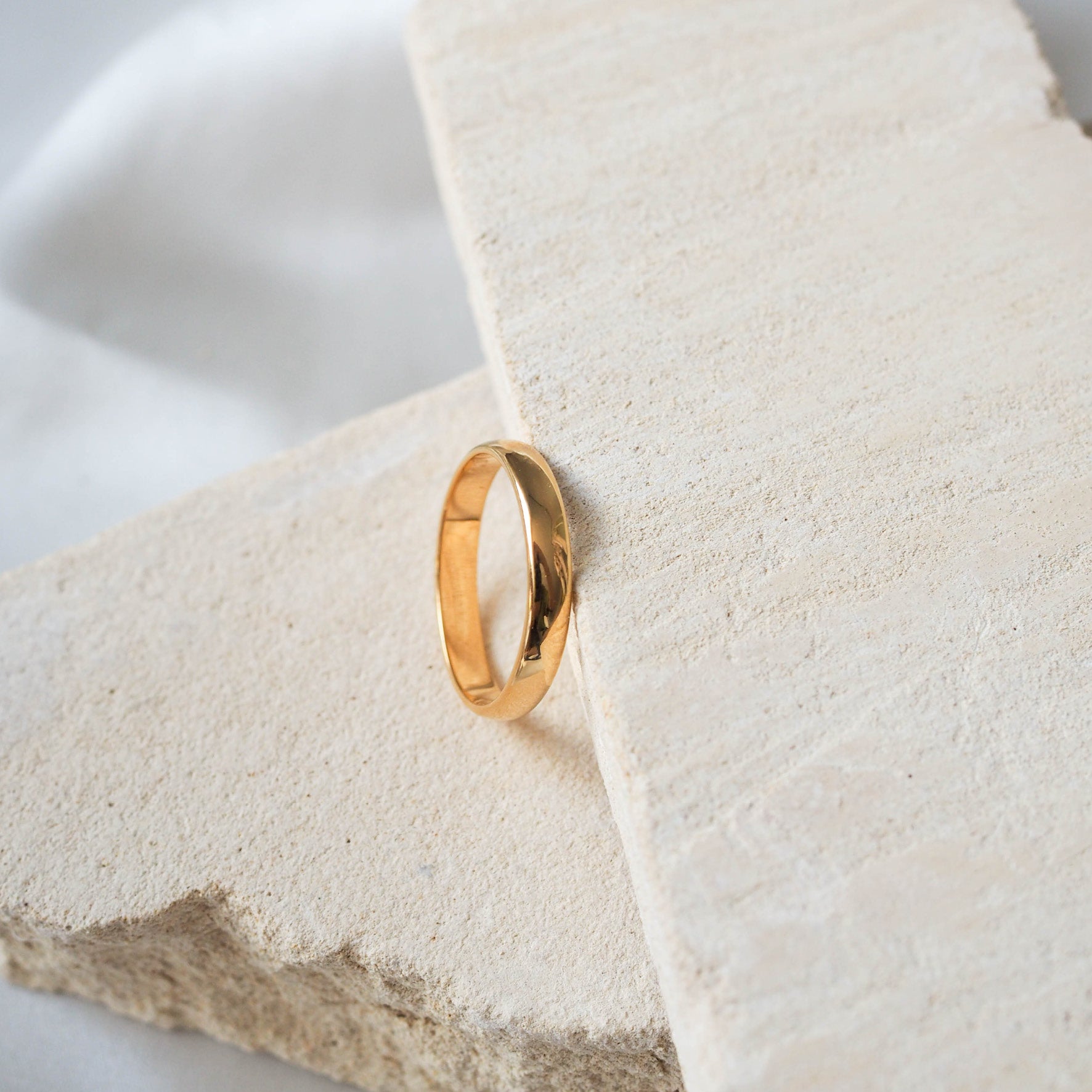 A single gold ring displayed on a textured beige stone, against a soft white background.