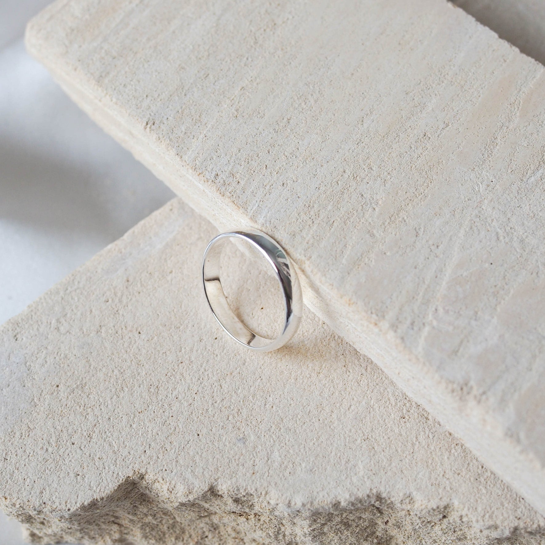 A single silver ring displayed on a textured beige stone, against a soft white background.
